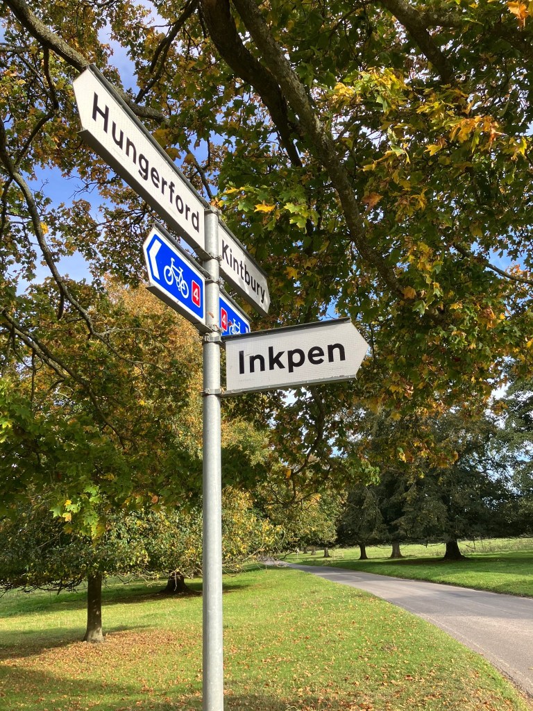 A fingerpost pointing to Inkpen, Hungerford and Kintbury. There are trees on a common with a road going through them behind the sign.